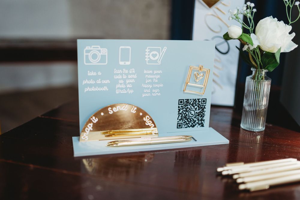 Elegant wedding sign with QR code, camera icons, and gold pen holder on wooden table with white floral arrangement.