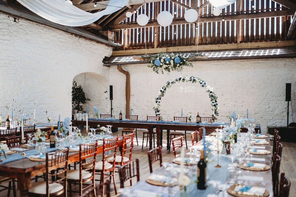 Elegant barn wedding reception with long wooden tables, blue linens, floral arch, white brick walls, and hanging paper lanterns. Tan y Graig