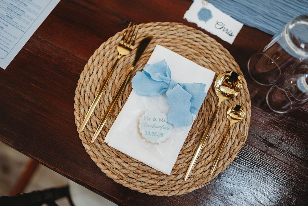 Elegant wedding place setting with woven charger, gold cutlery, blue bow menu card, and personalized cookie on wood table.
