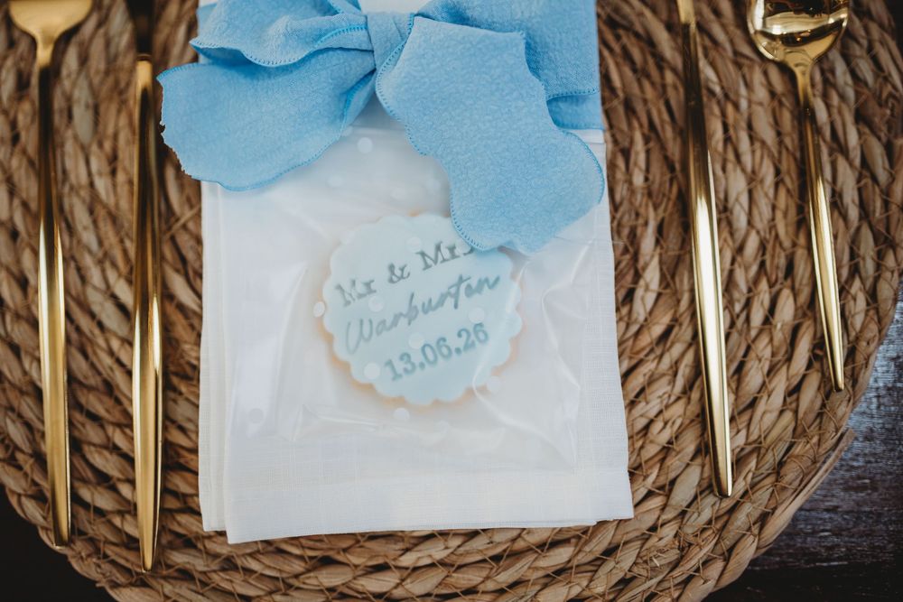 Elegant wedding place setting with gold cutlery, woven charger, blue ribbon, and personalized favor tag on white envelope.