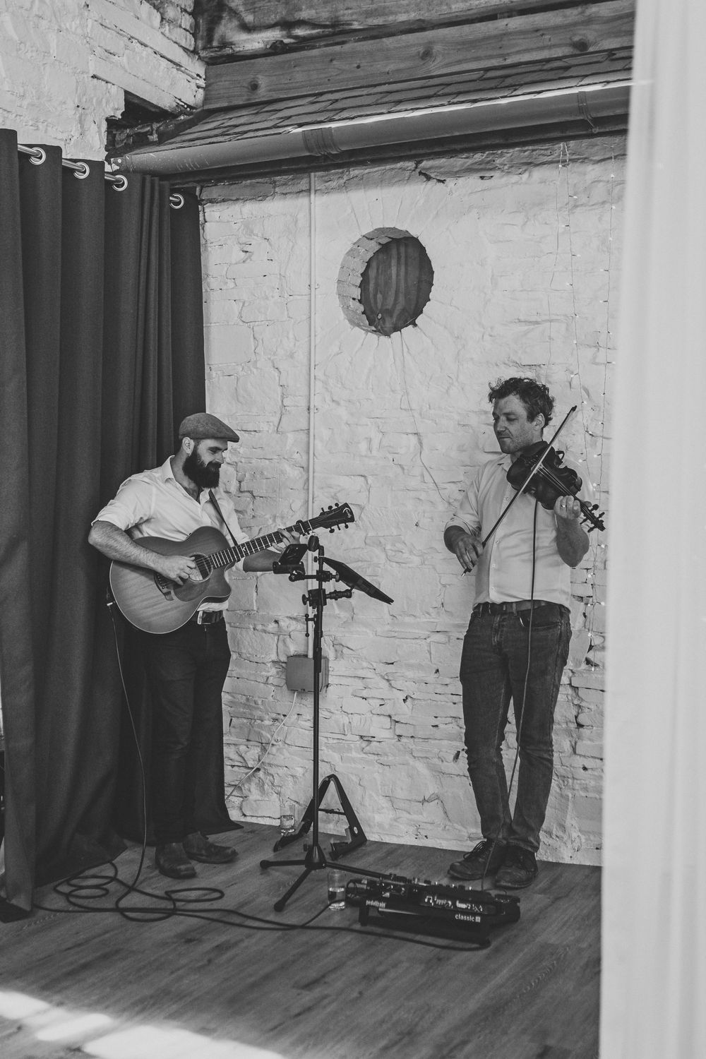 Two male musicians perform in a rustic brick room, one playing guitar and one playing violin, in black and white.
