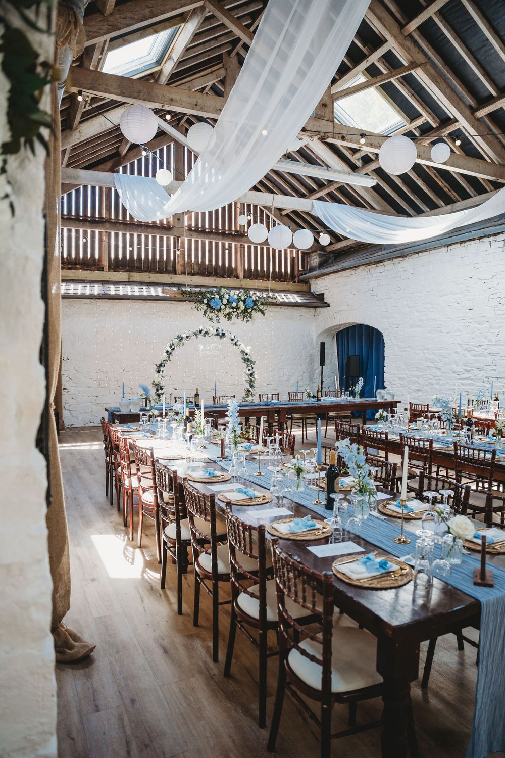 Rustic barn wedding reception with long wooden tables, white draping, floral arch, and globe lights in a whitewashed venue. Tan y Graig