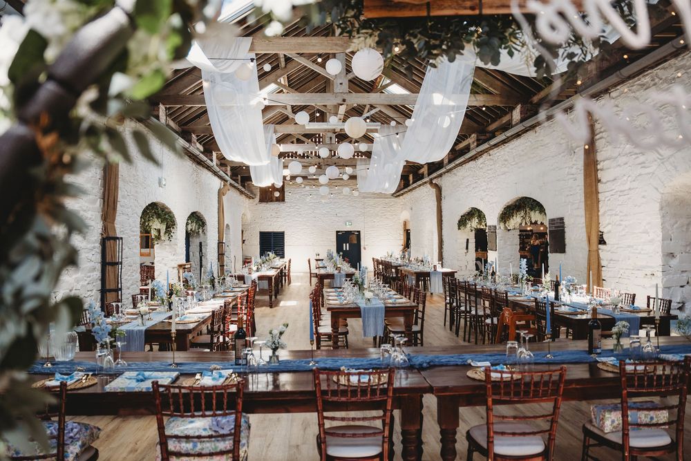 Elegant barn wedding reception with long tables, wooden chairs, white draping, and arched windows in a rustic white venue. Tan y Graig