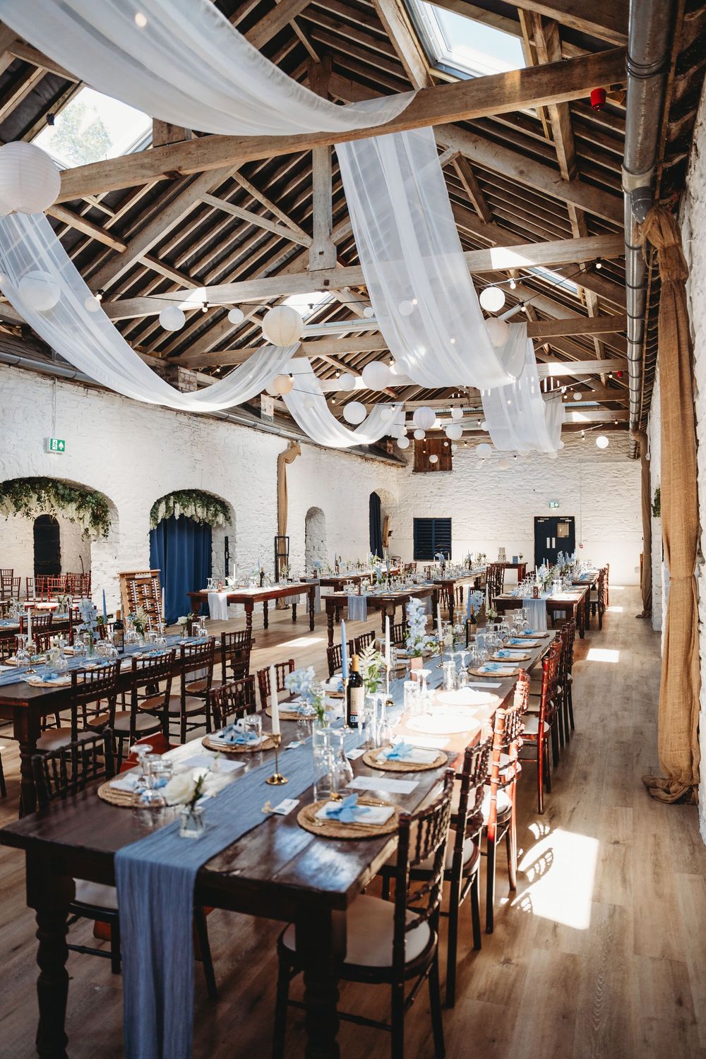 Rustic barn wedding reception with draped white fabric ceiling, long wooden tables, blue runners, and whitewashed brick walls. Tan y Graig