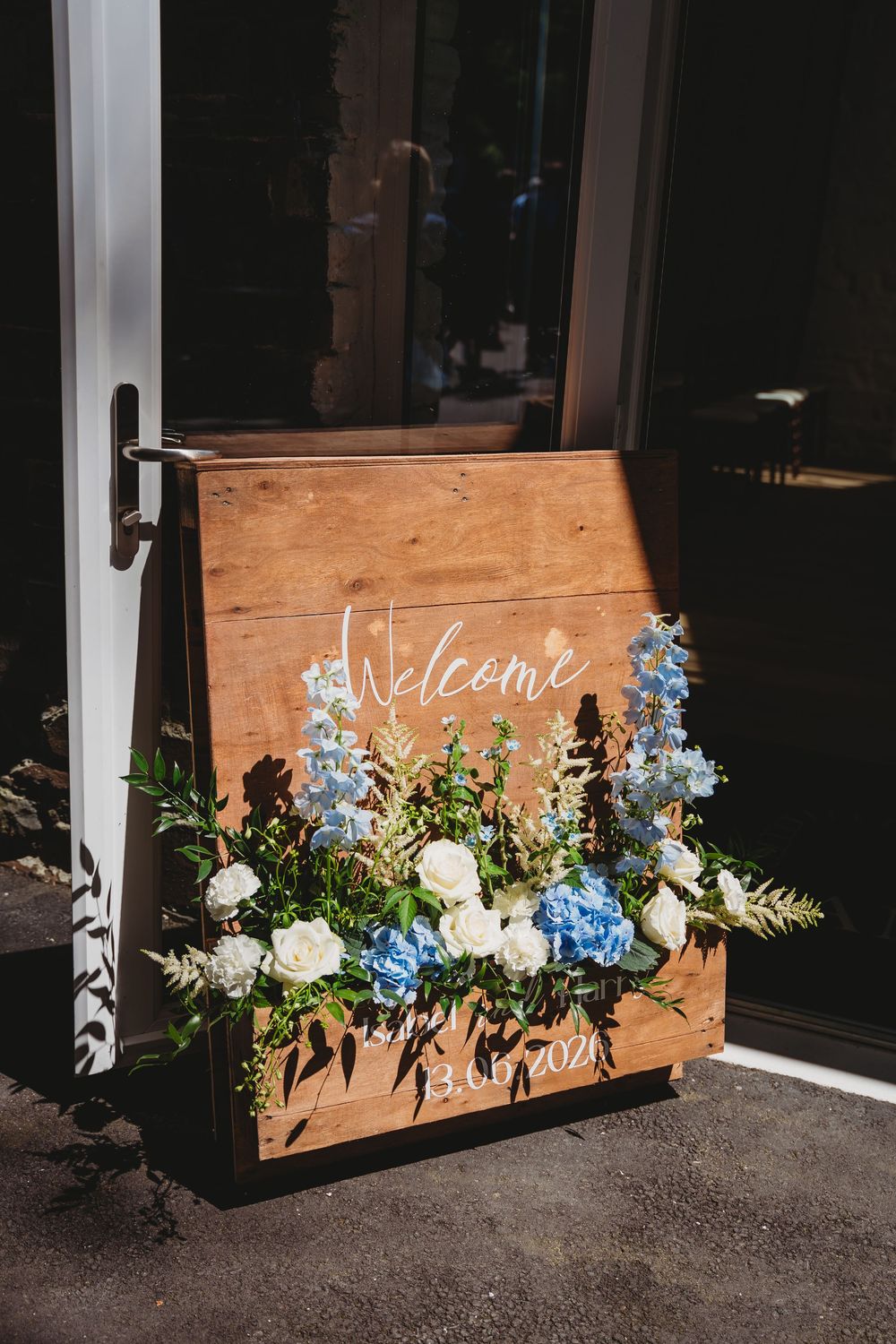Rustic wooden wedding welcome sign adorned with blue hydrangeas, white roses, and greenery, dated 13.6.2021.