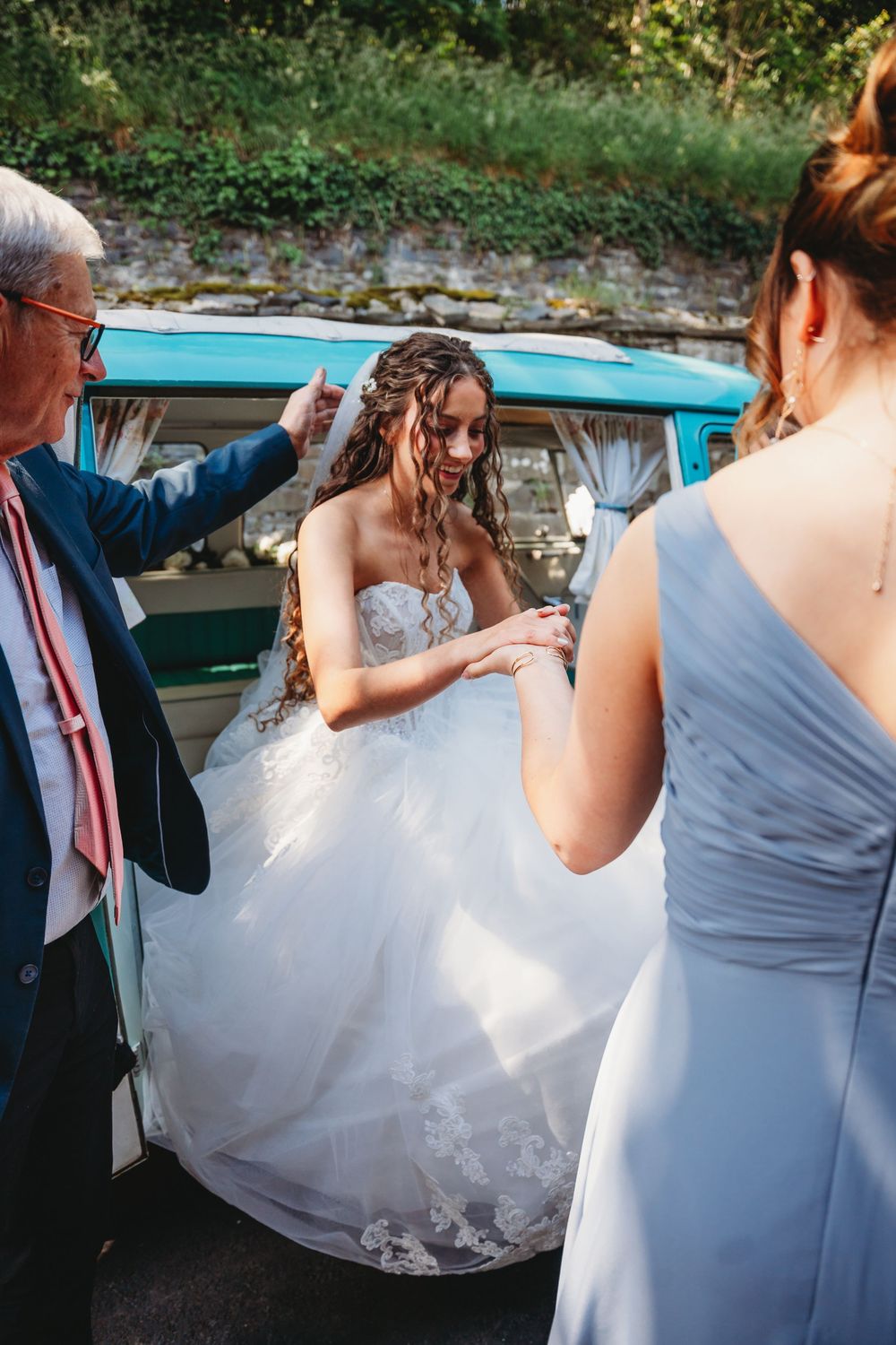 Bride in white lace gown exits vintage teal VW van assisted by guests on her wedding day.