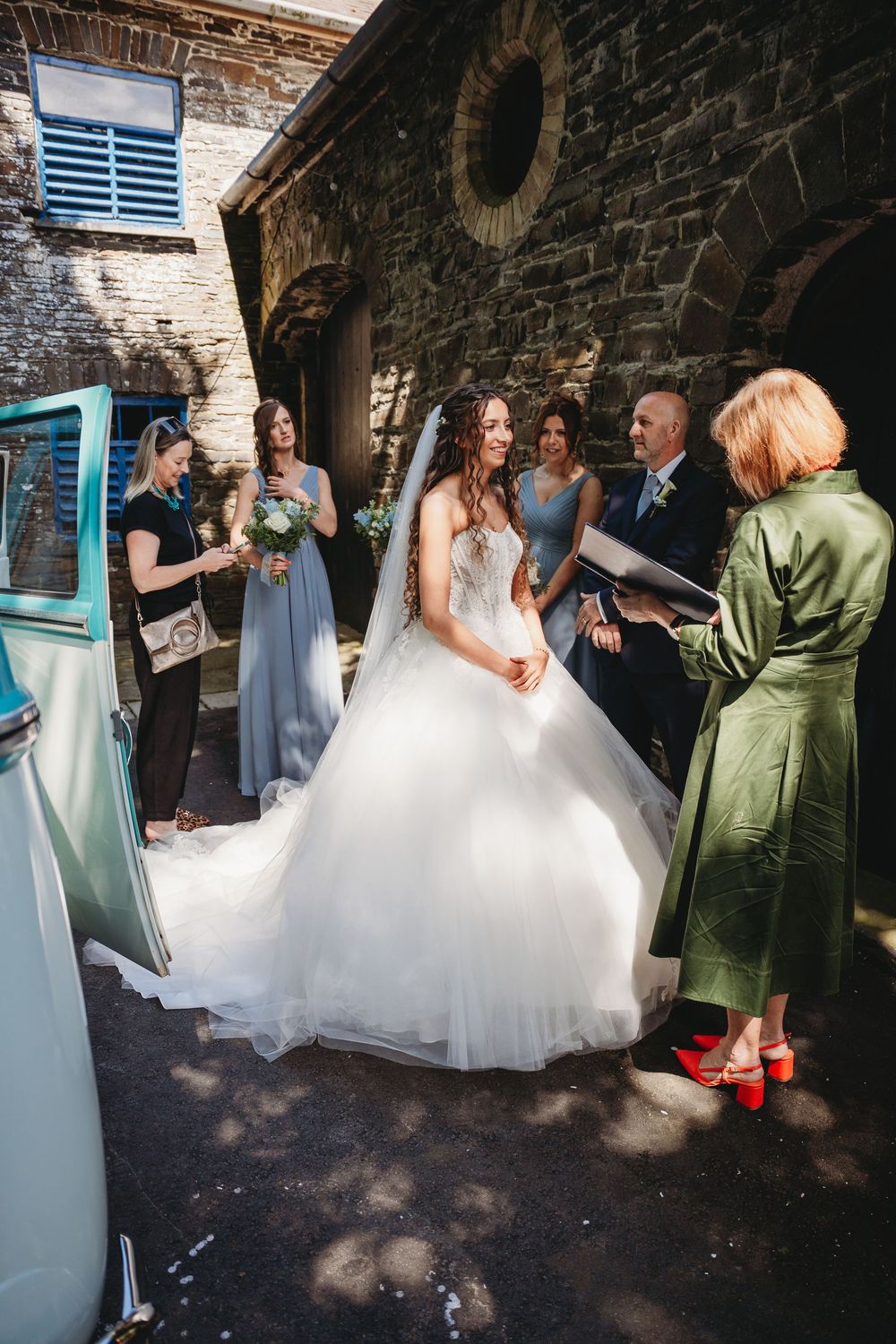 Bride in ballgown arrives at stone venue with bridesmaids in blue dresses and guests outdoors.
