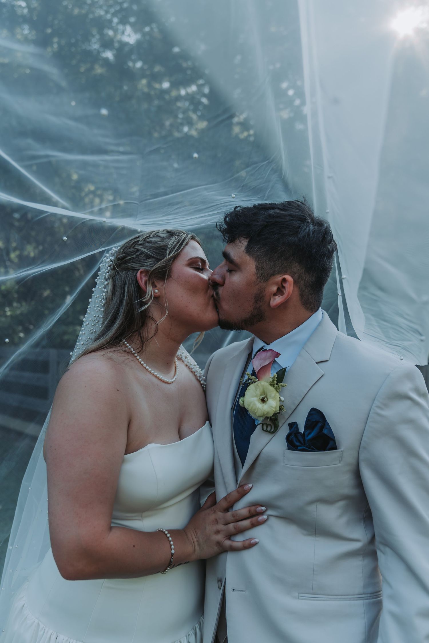 Bride and groom share a romantic kiss under a flowing white veil outdoors on their wedding day.