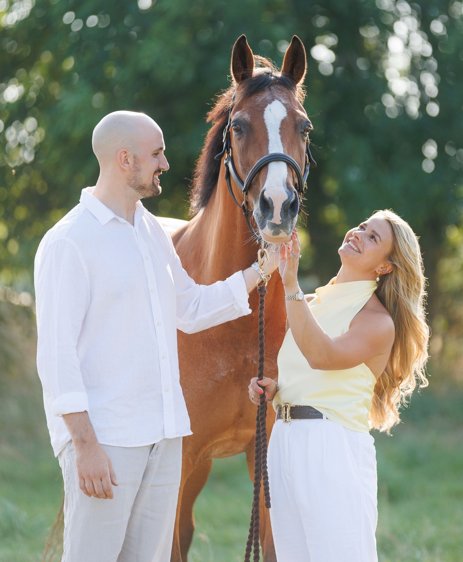 Woman in white lace dress walks alongside a brown horse on a sunlit forest path.