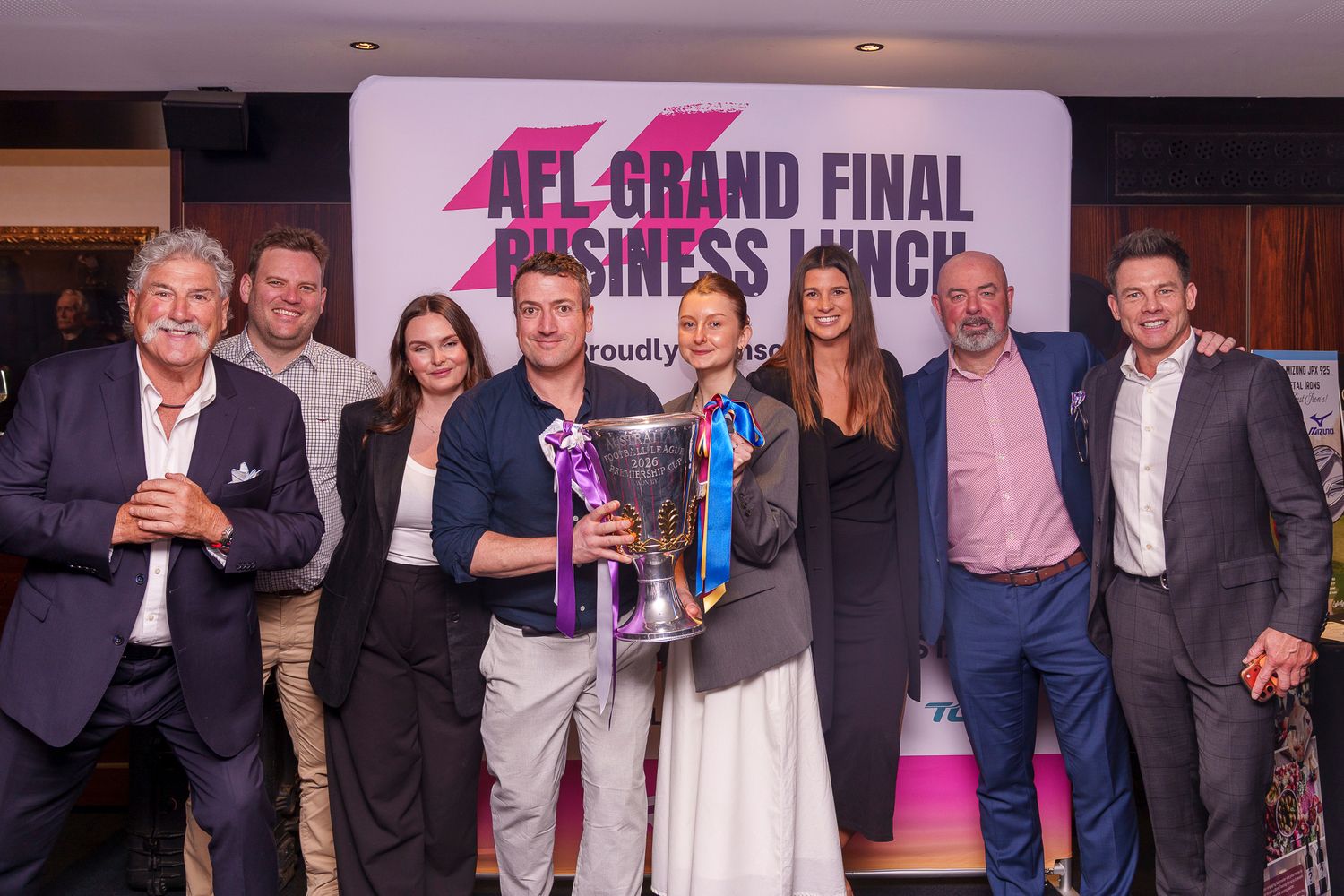 Group of professionals celebrating at AFL Grand Final Business Lunch event, holding trophy with purple ribbon.