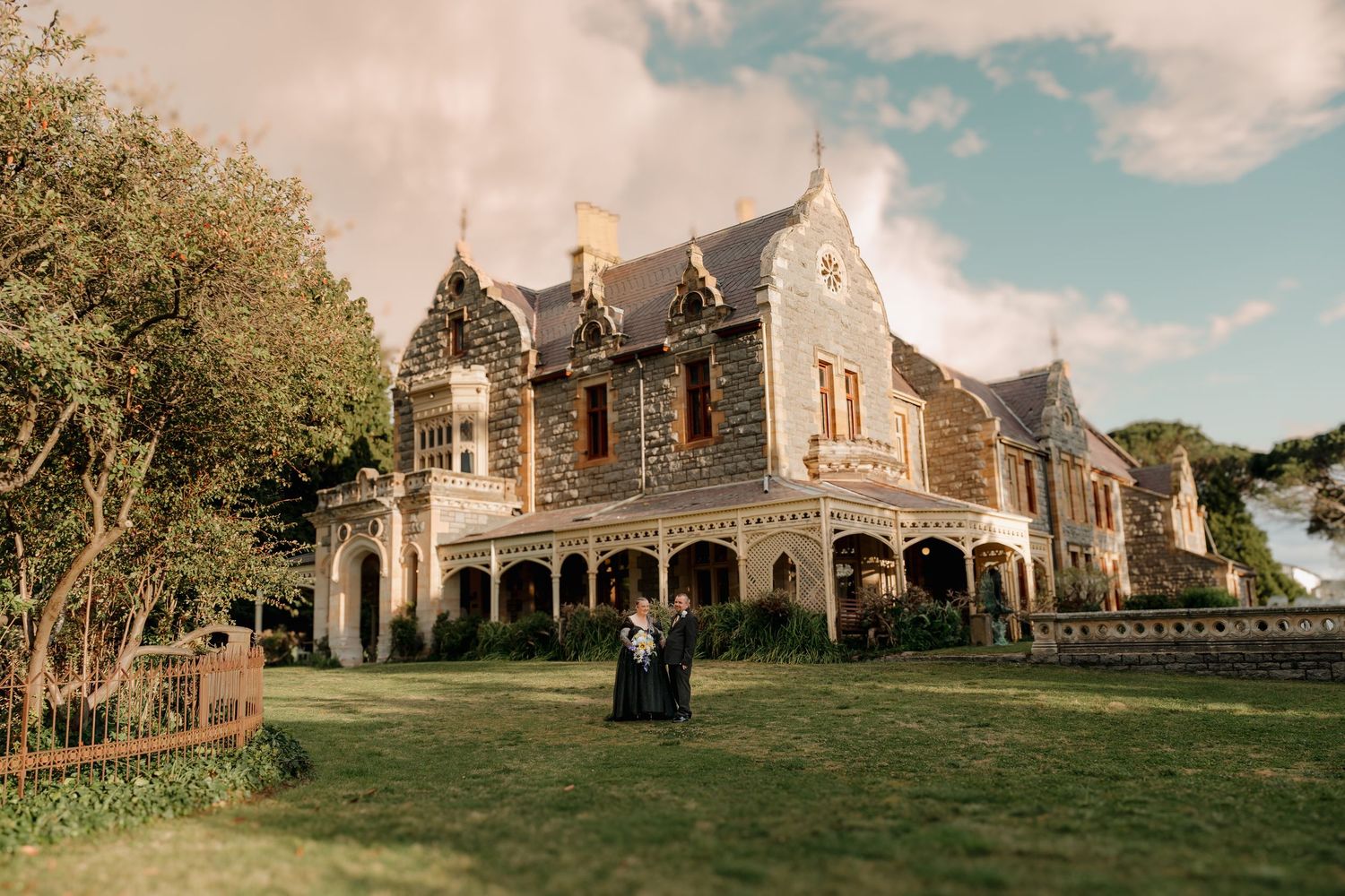 Couple in formal attire standing before a grand Victorian stone mansion with ornate architecture on lush green grounds.