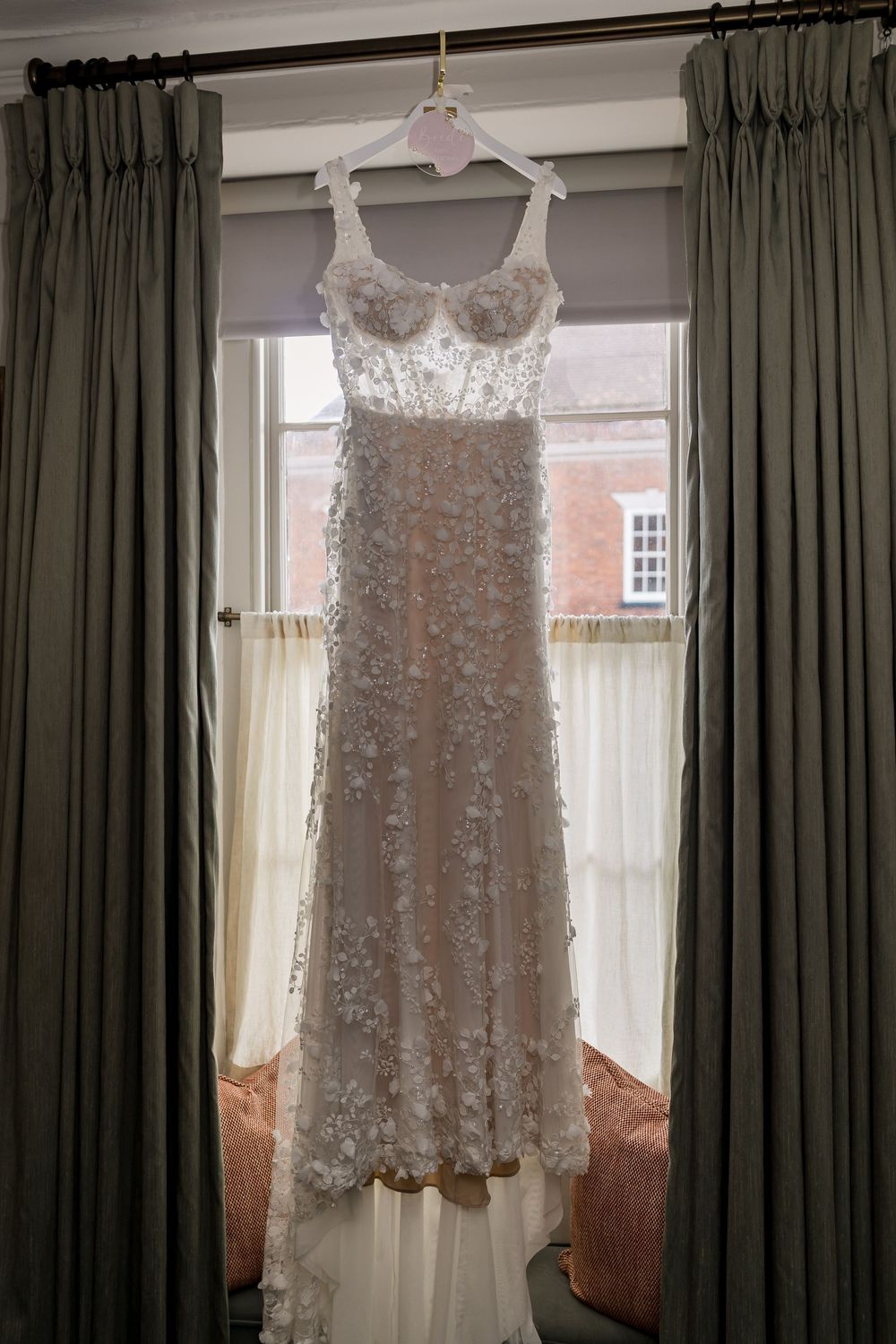 Elegant floral lace wedding dress hanging on a curtained window, featuring 3D flower appliques and a fitted silhouette.