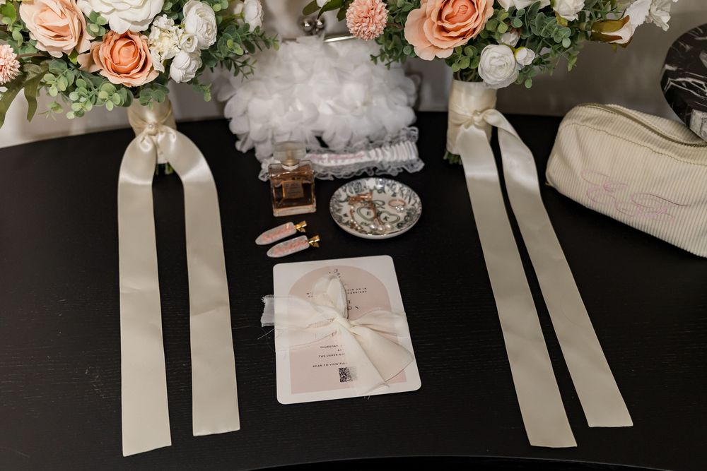Wedding accessories flat lay with floral arrangement, perfume, ribbon, jewelry dish, and white purse on dark surface.