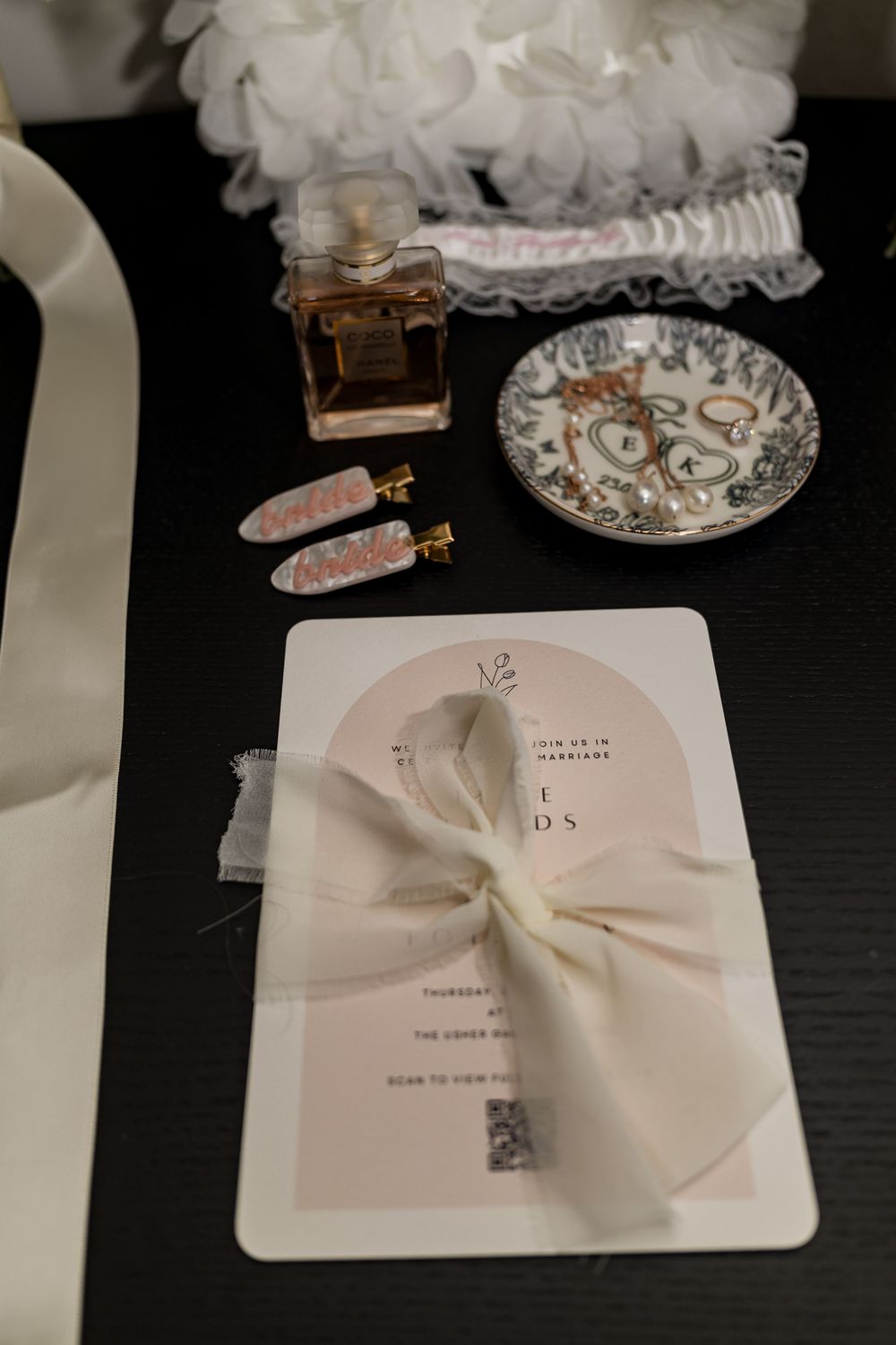Elegant wedding flat lay with invitation, ribbon bow, perfume bottle, garter, and jewelry dish on dark surface.