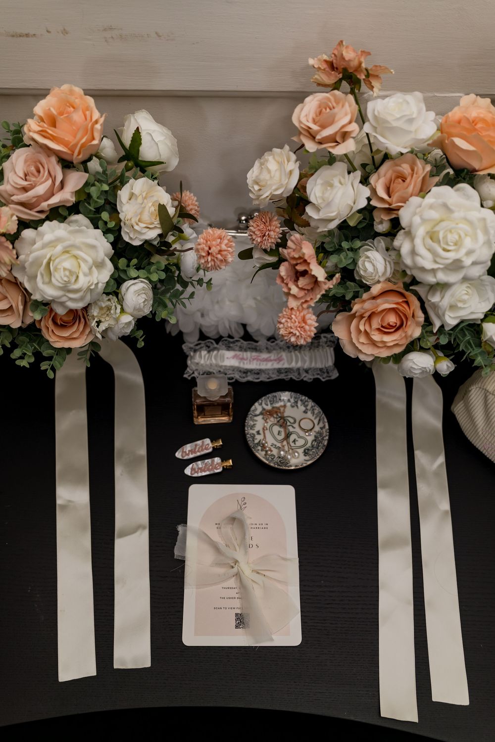 Elegant wedding flat lay with peach and white roses, ribbon accents, perfume, and stationery on a dark surface.