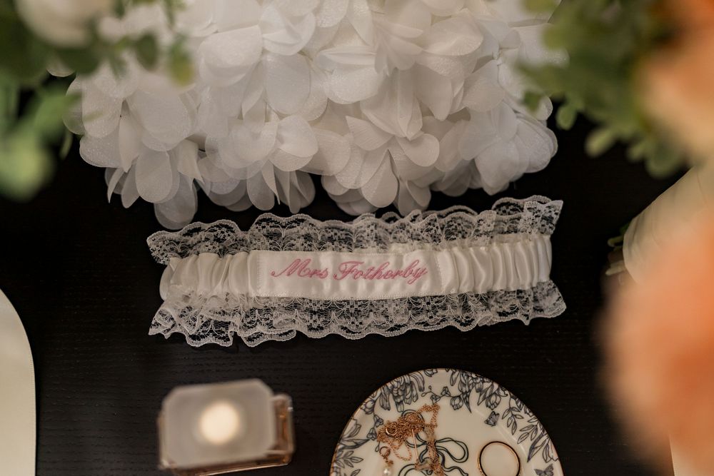 Personalized white lace wedding garter with pink embroidered name displayed on dark table with floral decor.