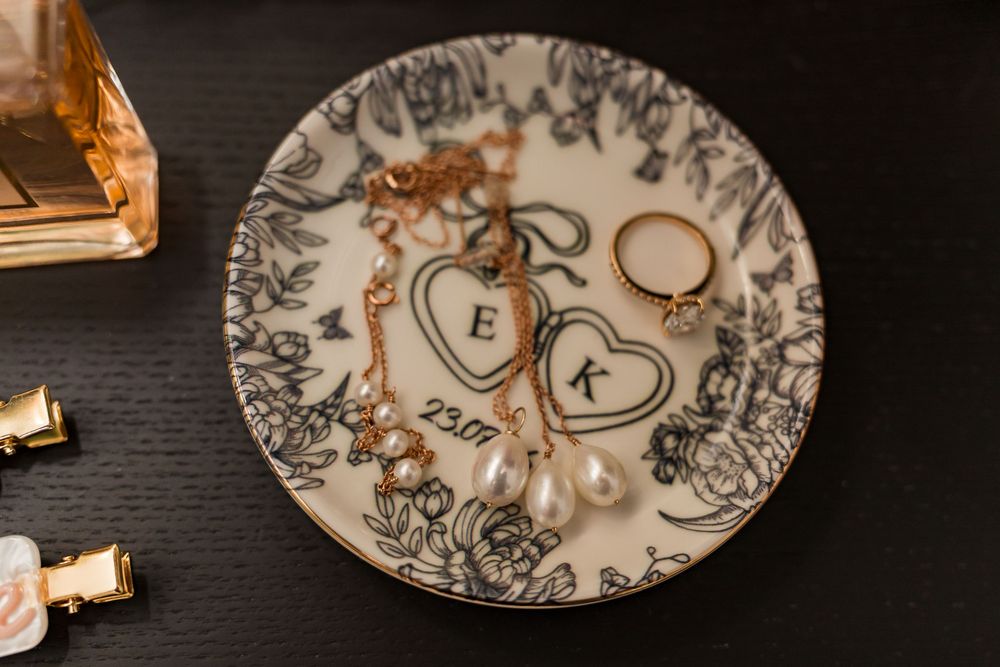 Gold jewelry and pearl earrings displayed on a floral monogram ring dish with initials E and K.