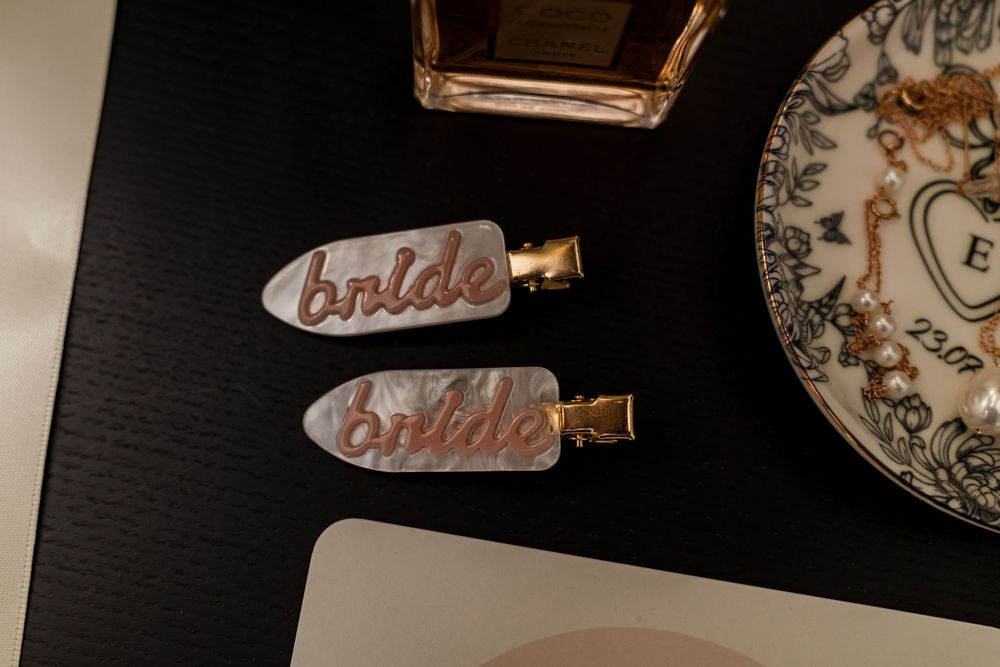 Two pearl bride hair clips with gold accents on a dark surface, next to a decorative plate and perfume bottle.