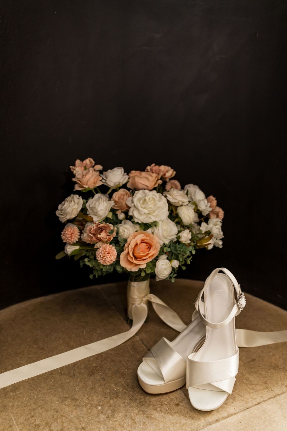 White bridal heels beside a bouquet of peach and white roses on a stone surface against a dark background.