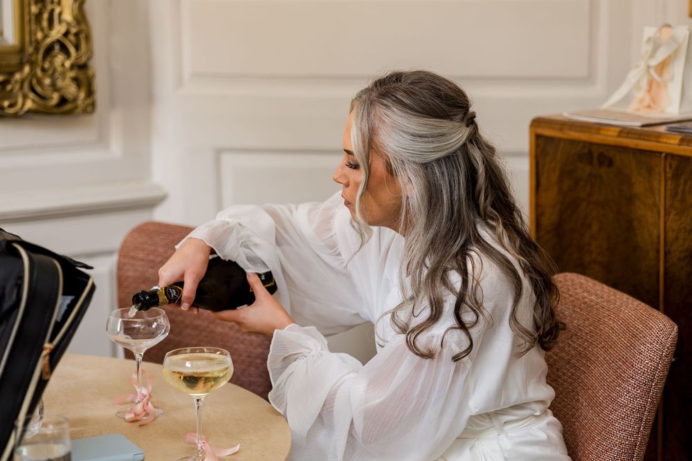 Woman with gray hair in white robe pouring champagne into glasses while sitting in elegant room.