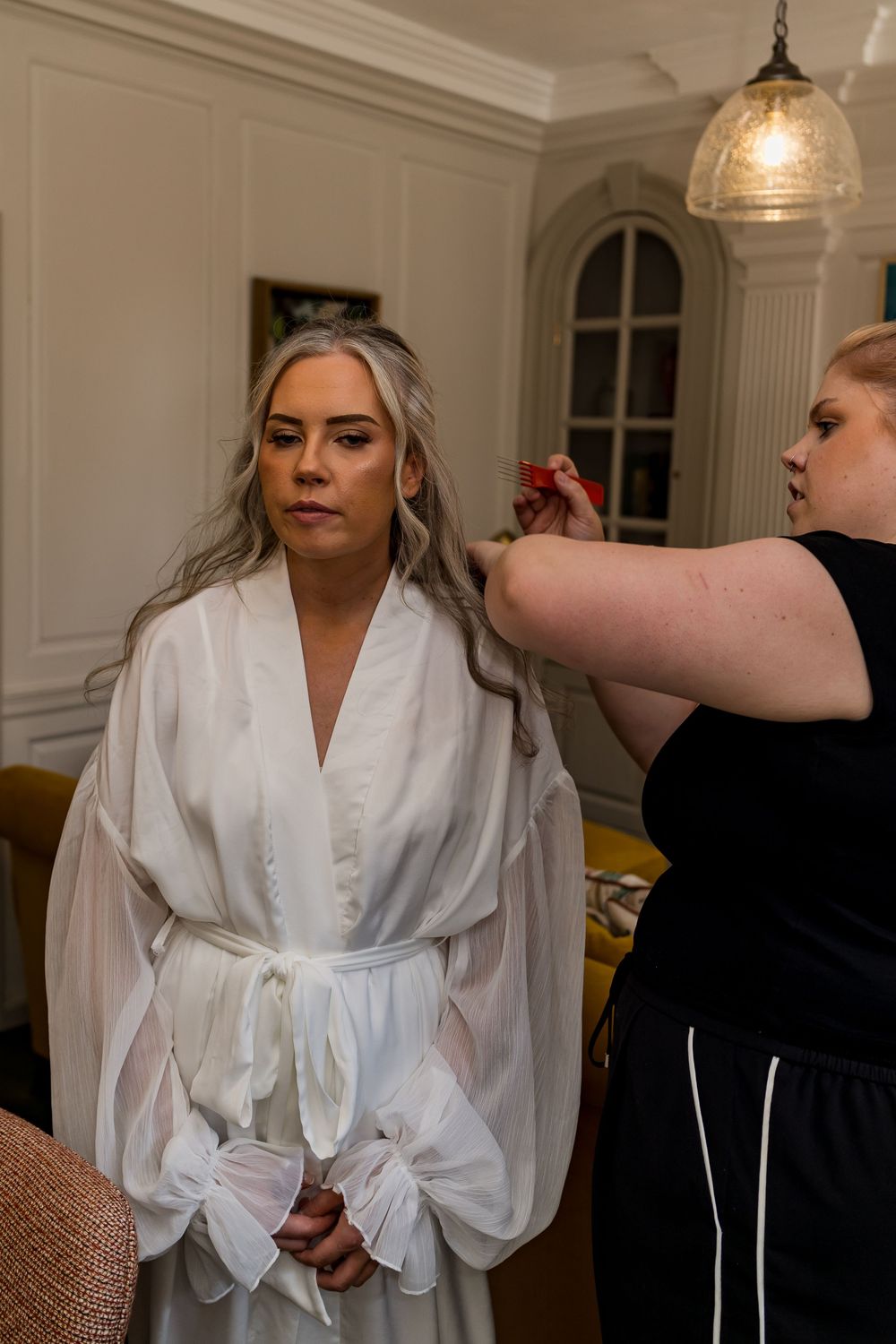 Bride in white robe getting hair styled by hairdresser during wedding morning preparations in elegant room.