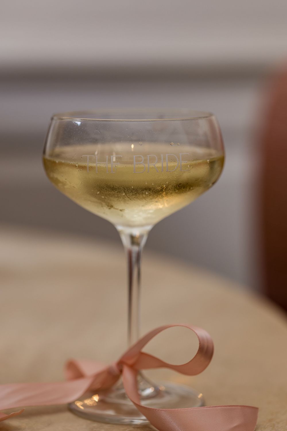 Elegant champagne coupe glass etched with text, filled with sparkling wine, adorned with a pink satin ribbon bow.