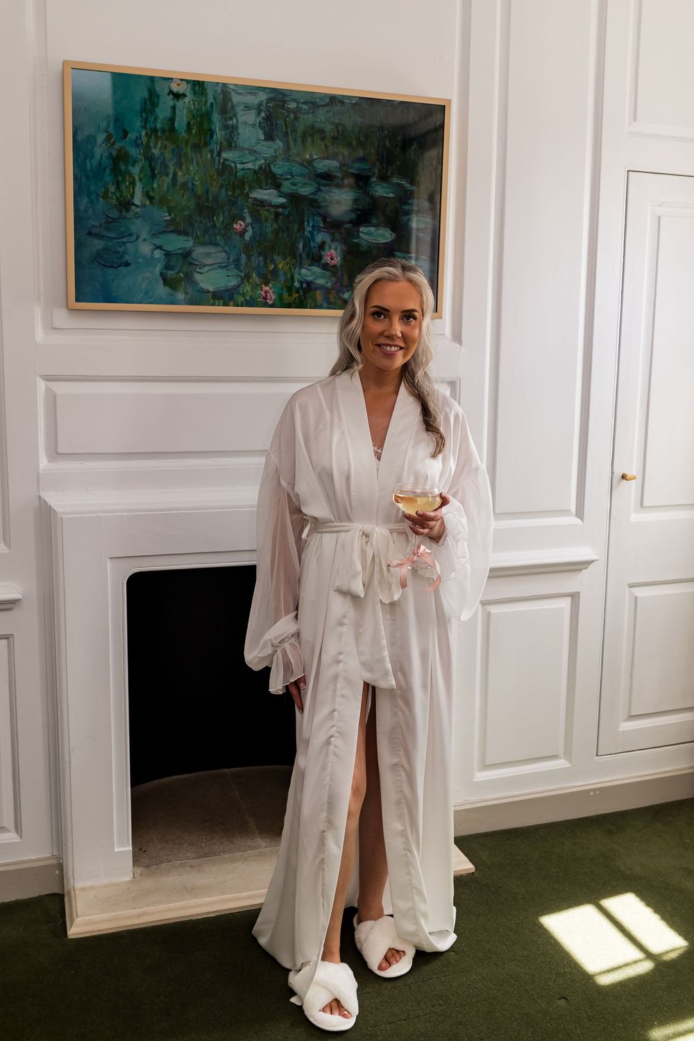 Woman in white robe holding champagne glass stands by fireplace with Monet water lilies painting.