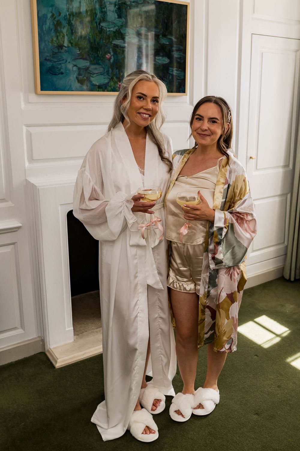 Two women in robes holding champagne glasses smiling together in elegant white-paneled room on wedding day.