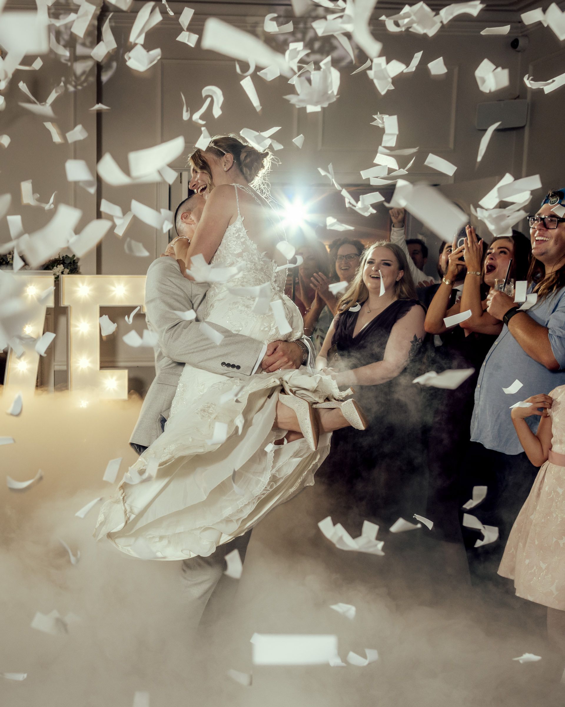 Bride and groom share a first dance as confetti floats through a misty reception while smiling wedding guests cheer in the background.