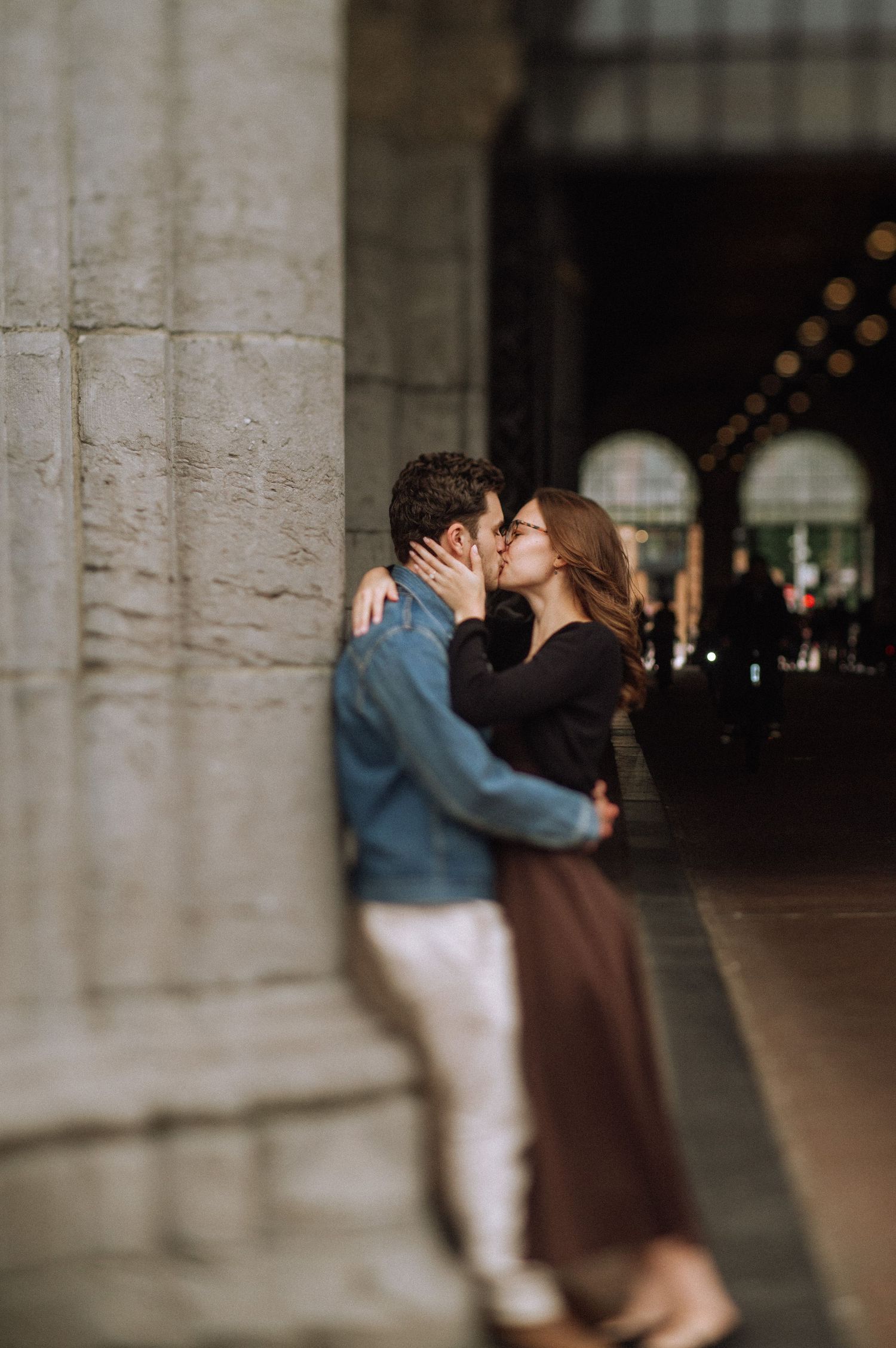 Couple kissing beside a large stone column in an arched passageway, the woman in a brown skirt and the man in a denim jacket.