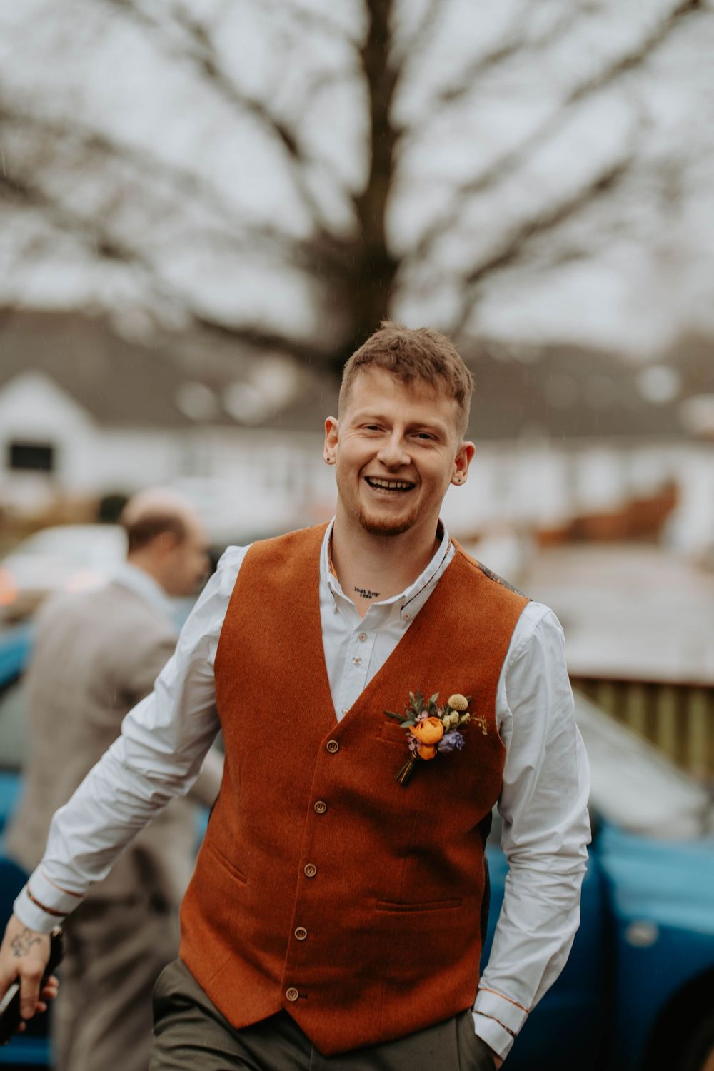 Smiling groom in rust orange vest with floral boutonniere laughing outdoors at wedding.