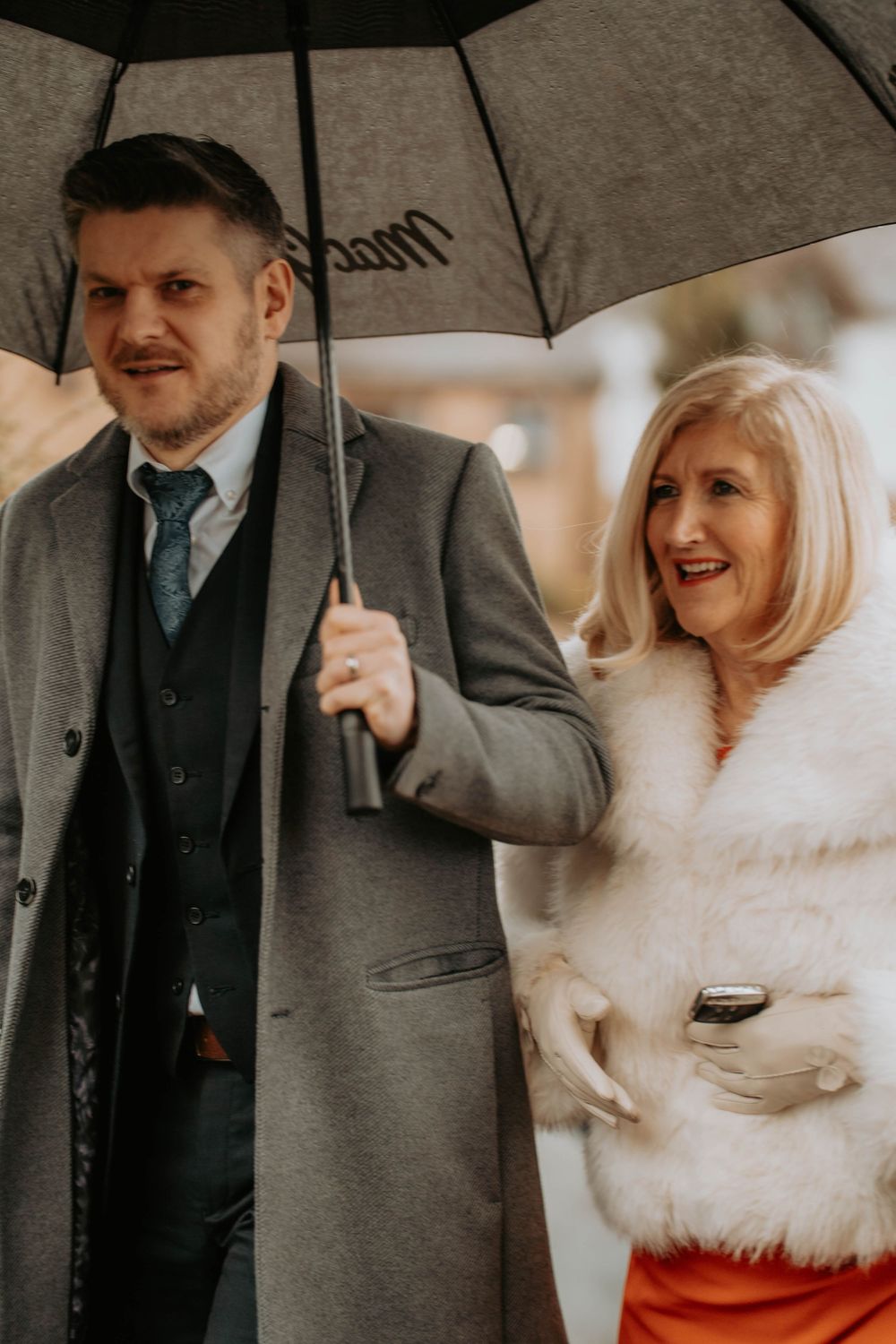 Couple sharing umbrella in rain, man in grey coat and tie, woman in white fur coat holding clutch bag.