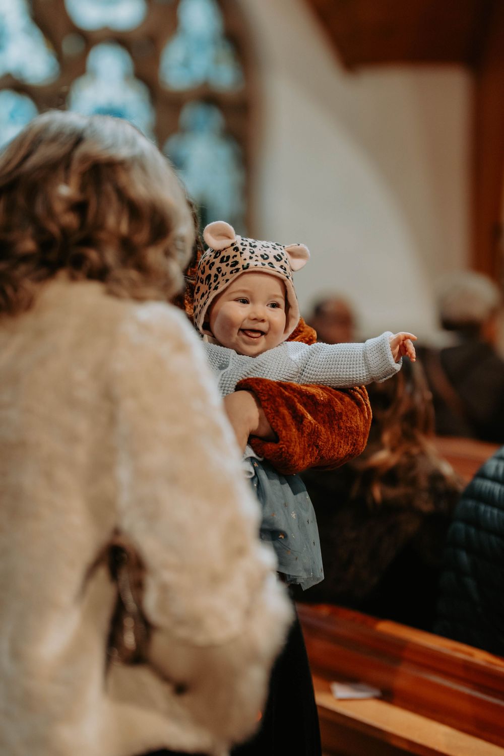 Smiling baby in a leopard print hat being held up in a church pew by a woman in a white fuzzy jacket.