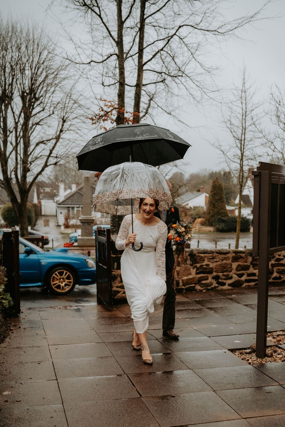 Bride in white dress walking in rain holding clear umbrella with bouquet on a wet pavement.