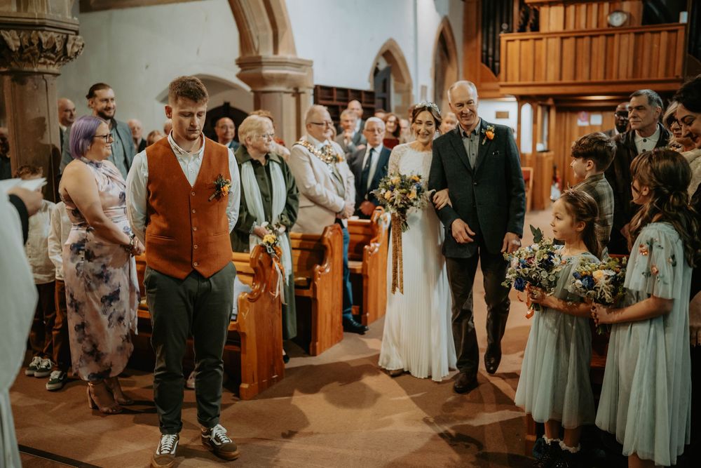 Bride walks down church aisle with escort as groom waits, guests watching in a rustic stone church wedding ceremony.