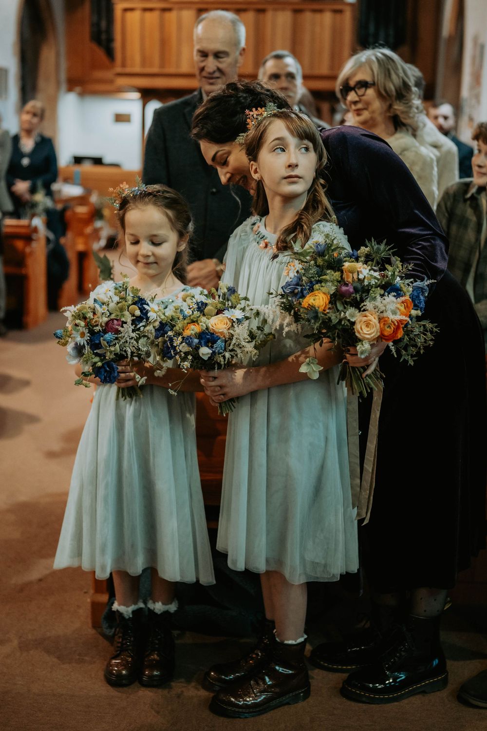 Two flower girls in pale blue dresses hold colorful bouquets at a church wedding ceremony.