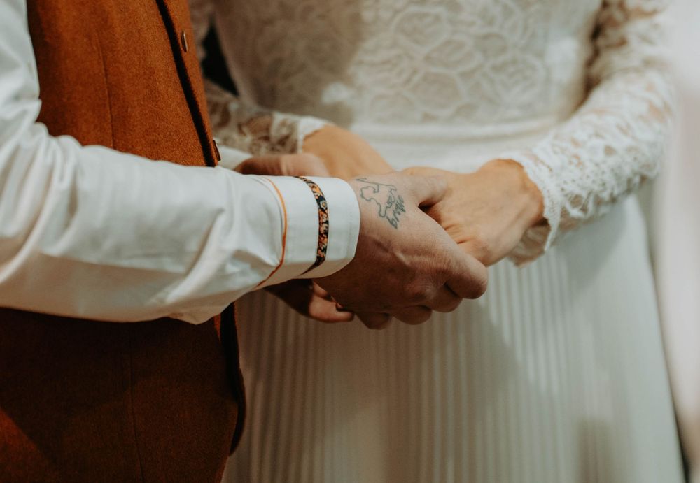 Bride and groom holding hands at wedding ceremony, groom wearing brown vest and bracelet, bride in lace white dress.