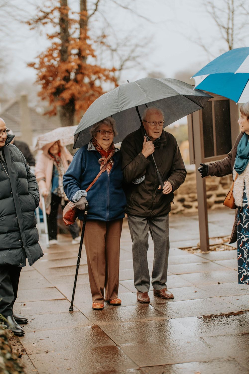 Elderly couple sharing an umbrella walking on a wet pavement in the rain at a wedding.