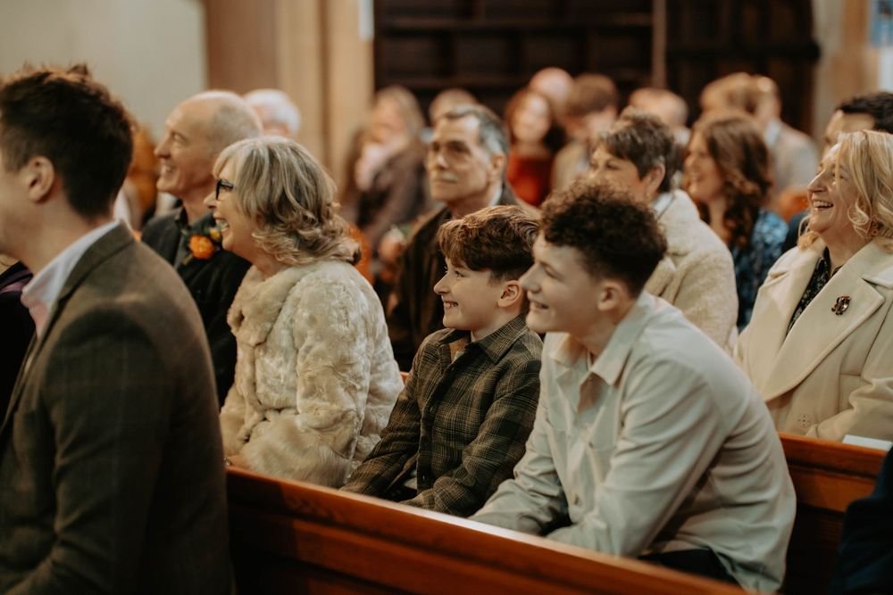 Guests smiling and seated in church pews during a wedding ceremony, dressed in smart casual attire.