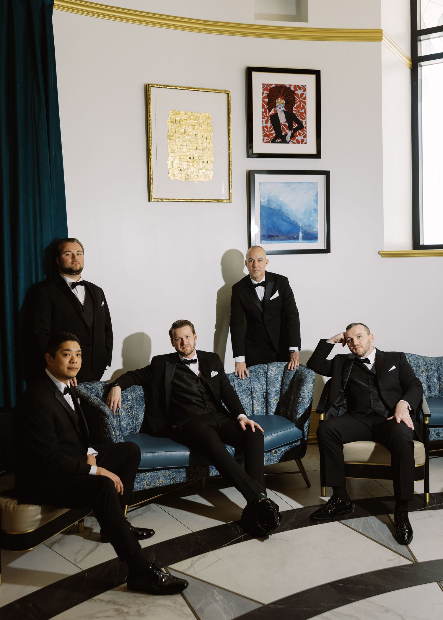 Five men in black tuxedos and bow ties pose elegantly in a stylish room with blue velvet furniture and framed artwork.