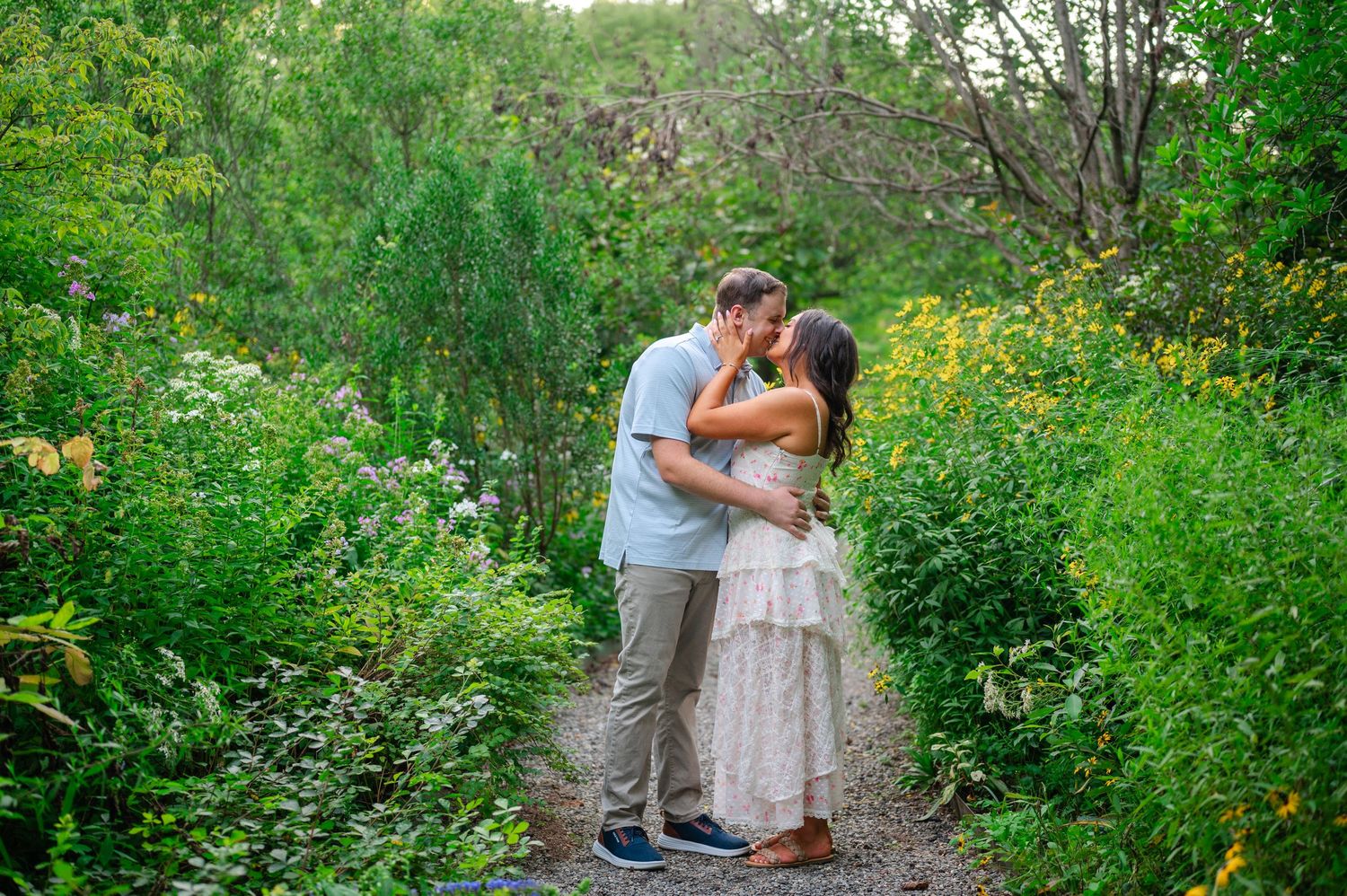 Couple sharing a romantic kiss on a garden path surrounded by lush green foliage and wildflowers in Cross Estate Gardens in NJ.