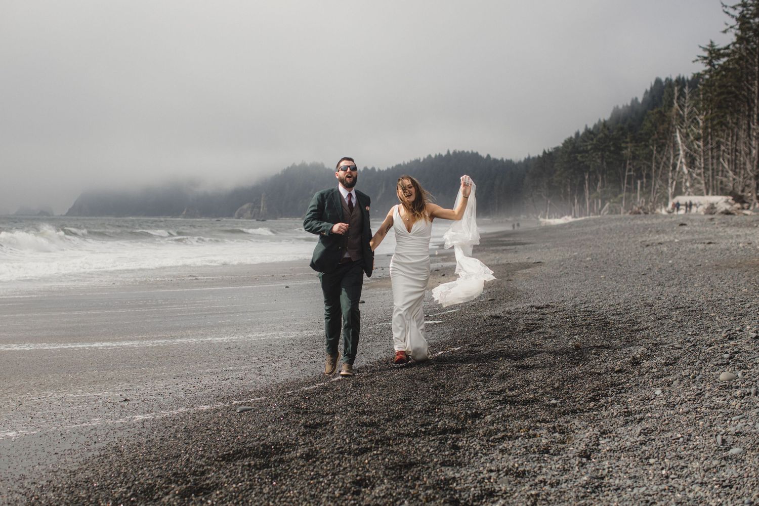 Bride and groom walk hand-in-hand on a misty Pacific Northwest beach, veil flowing in the wind.