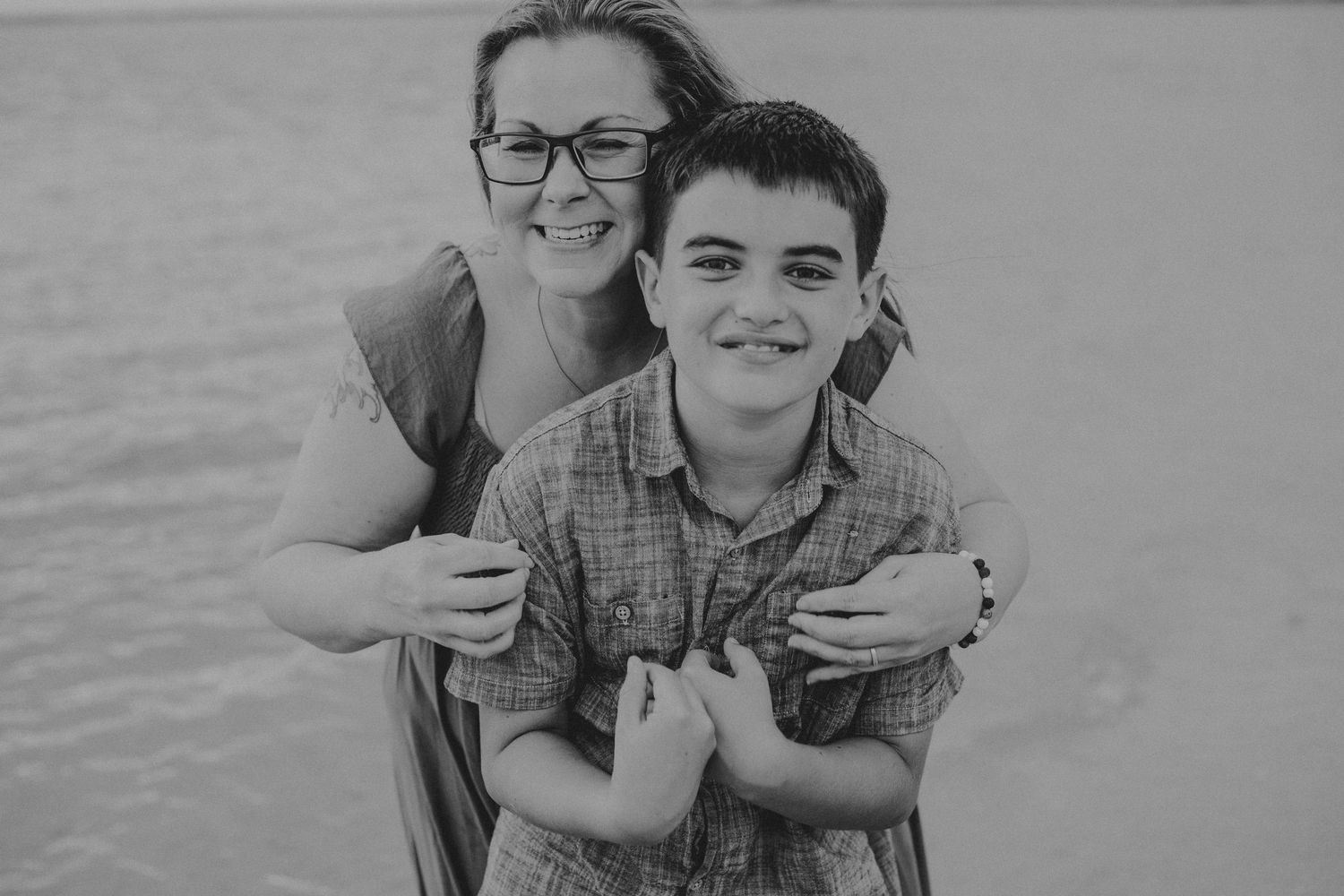 Black and white photo of a smiling woman with glasses hugging a happy boy in a plaid shirt from behind at the beach.