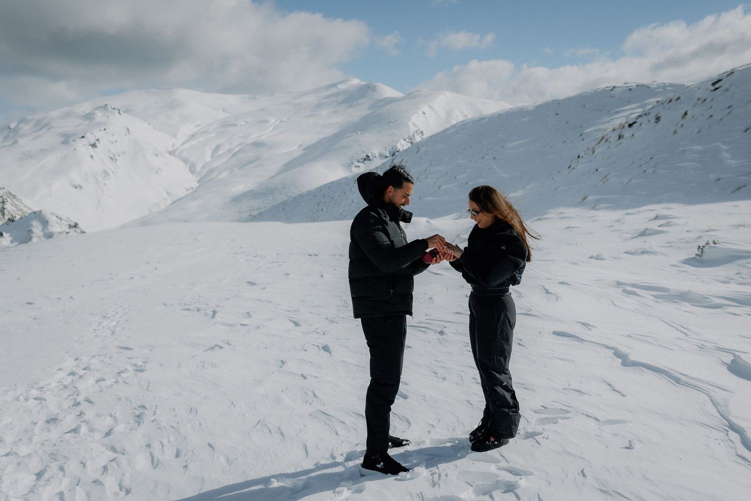 Couple holding hands on a snowy mountain landscape with white peaks and blue sky in the background.