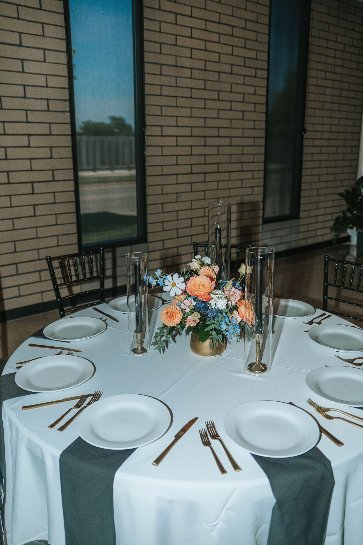 Elegant round wedding reception table with white linen, gold cutlery, white plates, and a peach and blue floral centerpiece.
