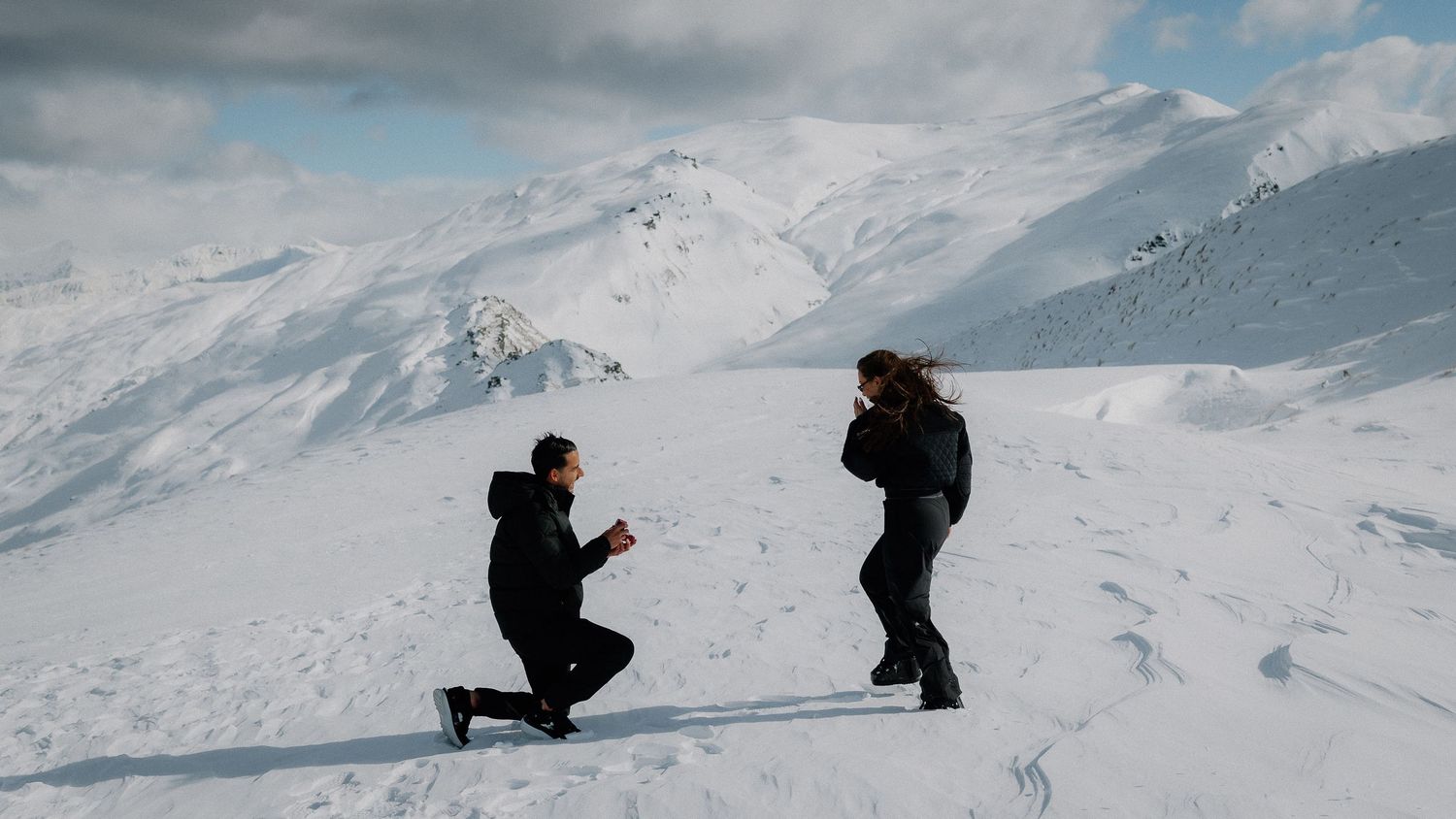Man proposes on snowy mountain top, kneeling before woman with stunning snow-capped peaks in background.