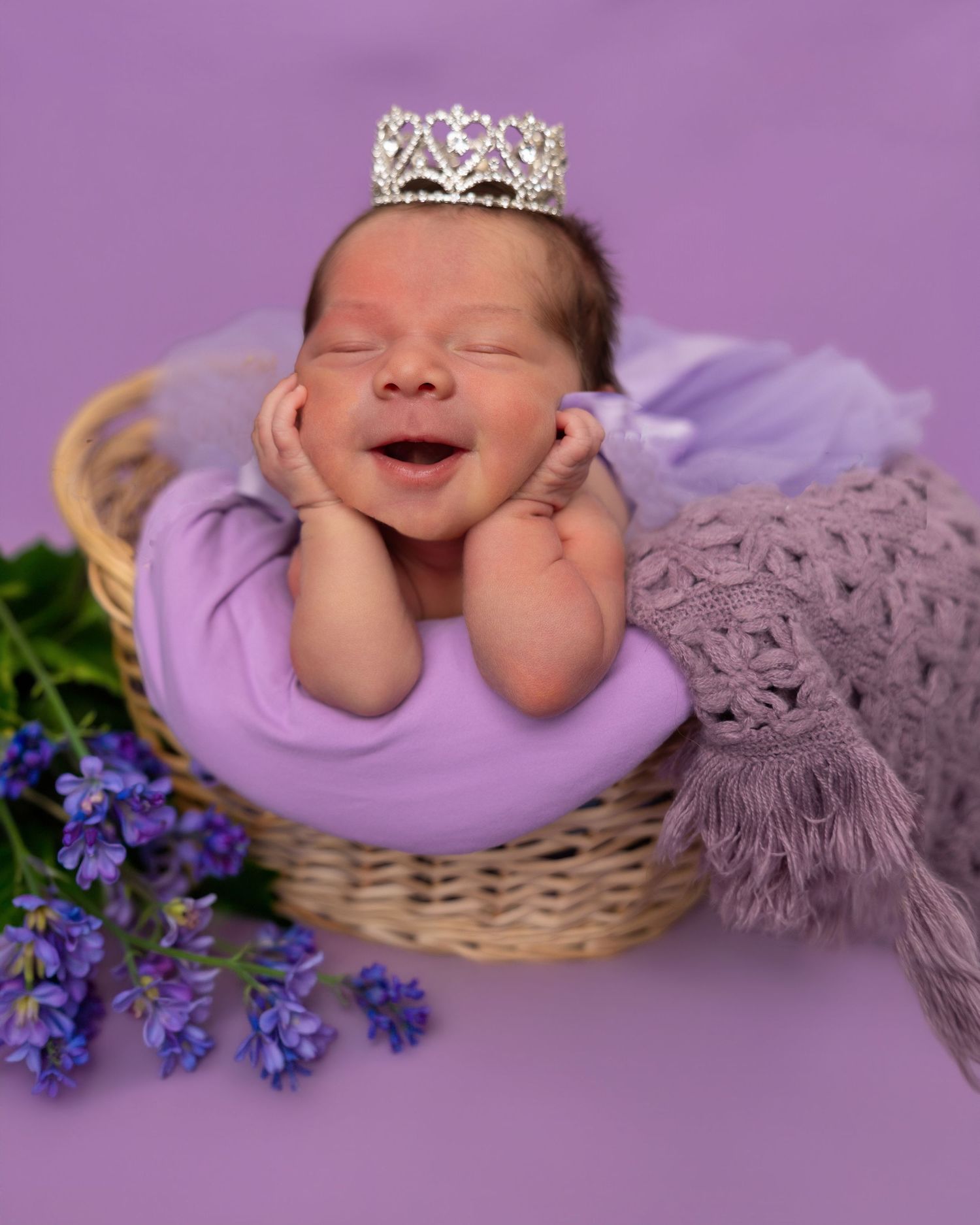Smiling newborn baby wearing a tiny silver crown, wrapped in purple, resting in a wicker basket with lavender flowers.