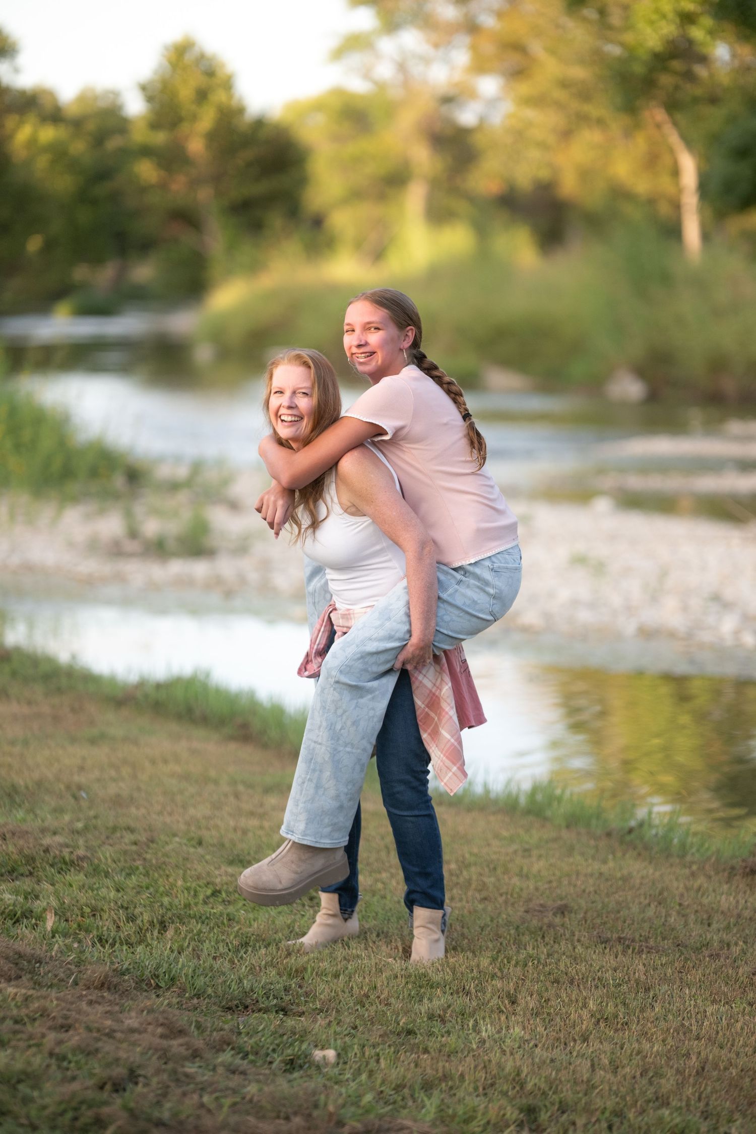 Two girls laughing outdoors near a river, one giving the other a piggyback ride on a sunny day.