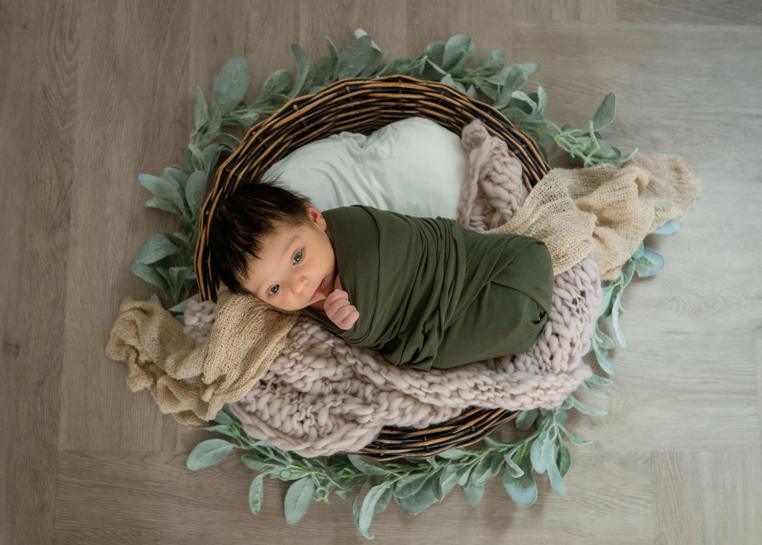 Newborn baby swaddled in green wrap sleeping in wicker basket with teddy bear and eucalyptus leaves.
