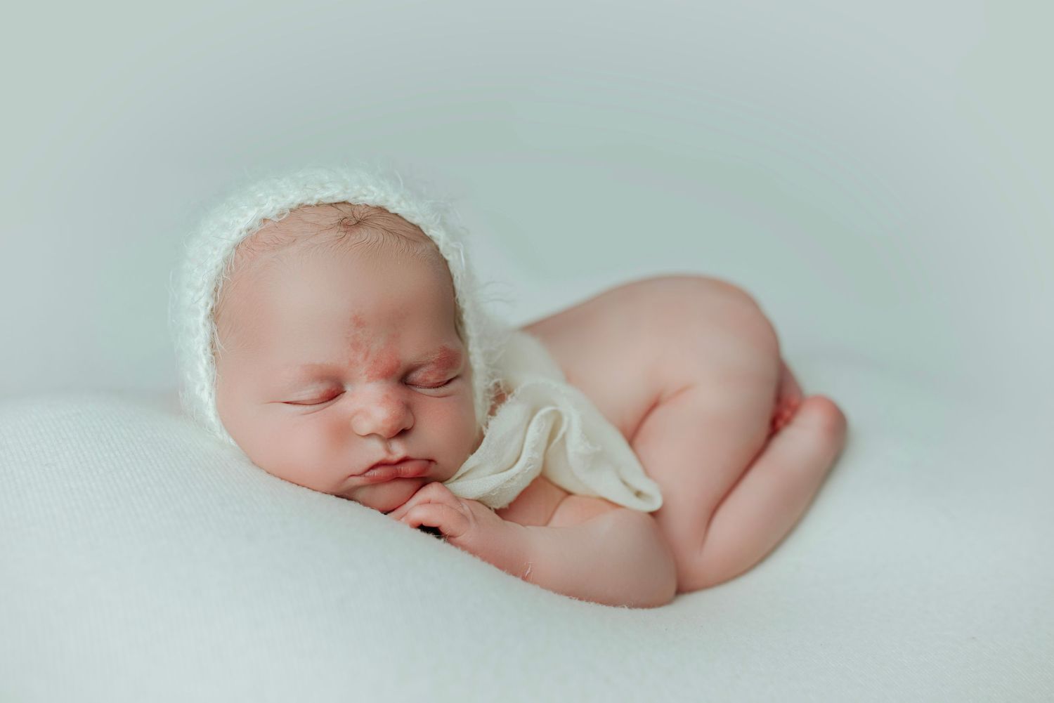 Newborn baby sleeping peacefully in a white bonnet and wrap on a soft light background.